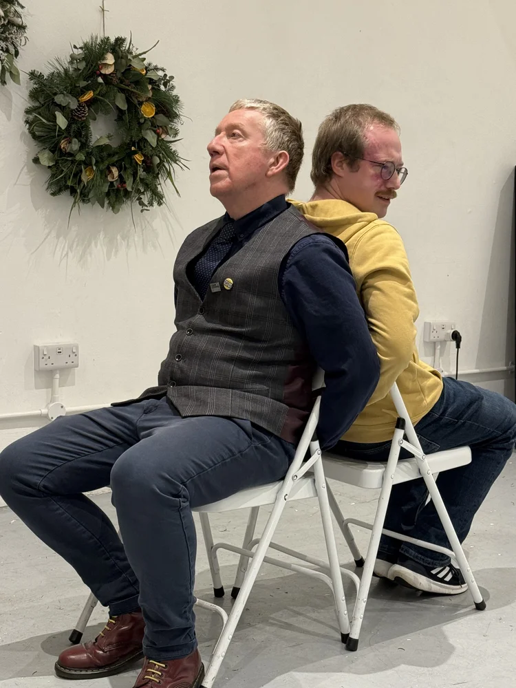East Midlands improvisers Adam Hodgson and Chris Collins are pictured sitting down back to back on two white chairs as if they have been taken hostage.