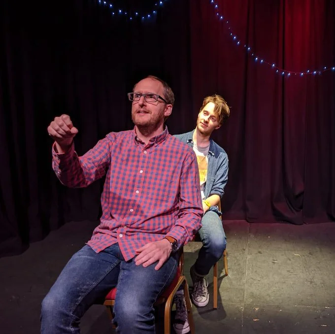 A picture of Nottingham improvisers Lloydie James Lloyd and Liam Webber. Lloydie sits in front of Liam, miming driving a car while Liam looks quizzically over his shoulder